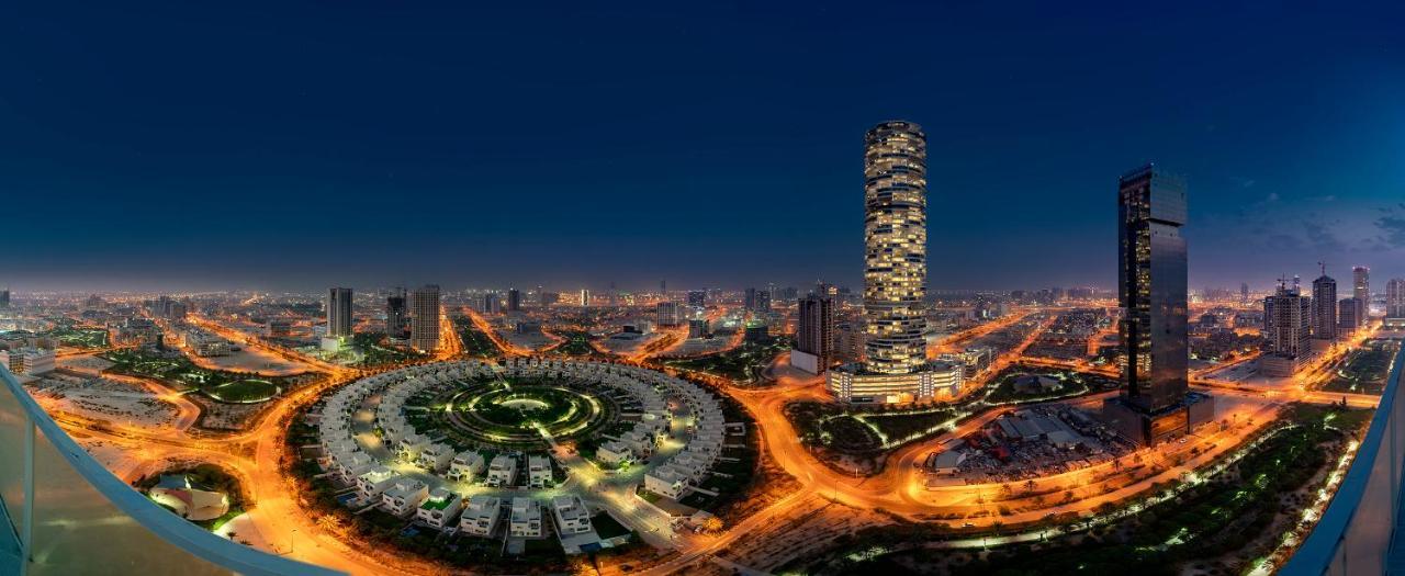 Five Jumeirah Village Дубай Экстерьер фото The photo showcases a cityscape illuminated at night. In the foreground, there is a circular arrangement of buildings or houses, creating a visually striking pattern. To the right, a tall, modern skyscraper stands out, featuring a sleek design with n