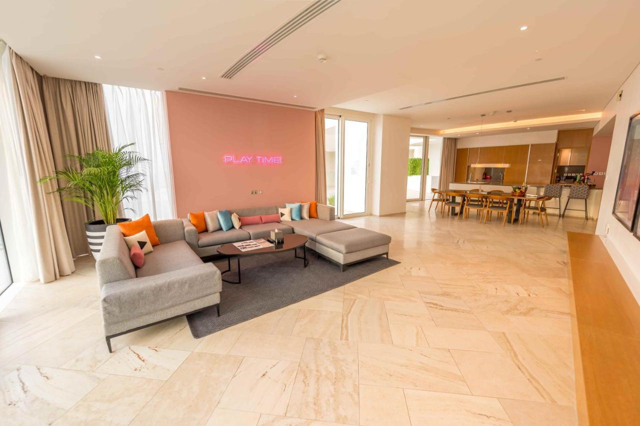 Five Jumeirah Village Дубай Экстерьер фото The photo shows a spacious and modern living area. There is a large, light-colored sectional sofa with colorful cushions, positioned on a dark rug. The wall behind the sofa is painted in a soft pink shade, and there is a subtle decorative element or 