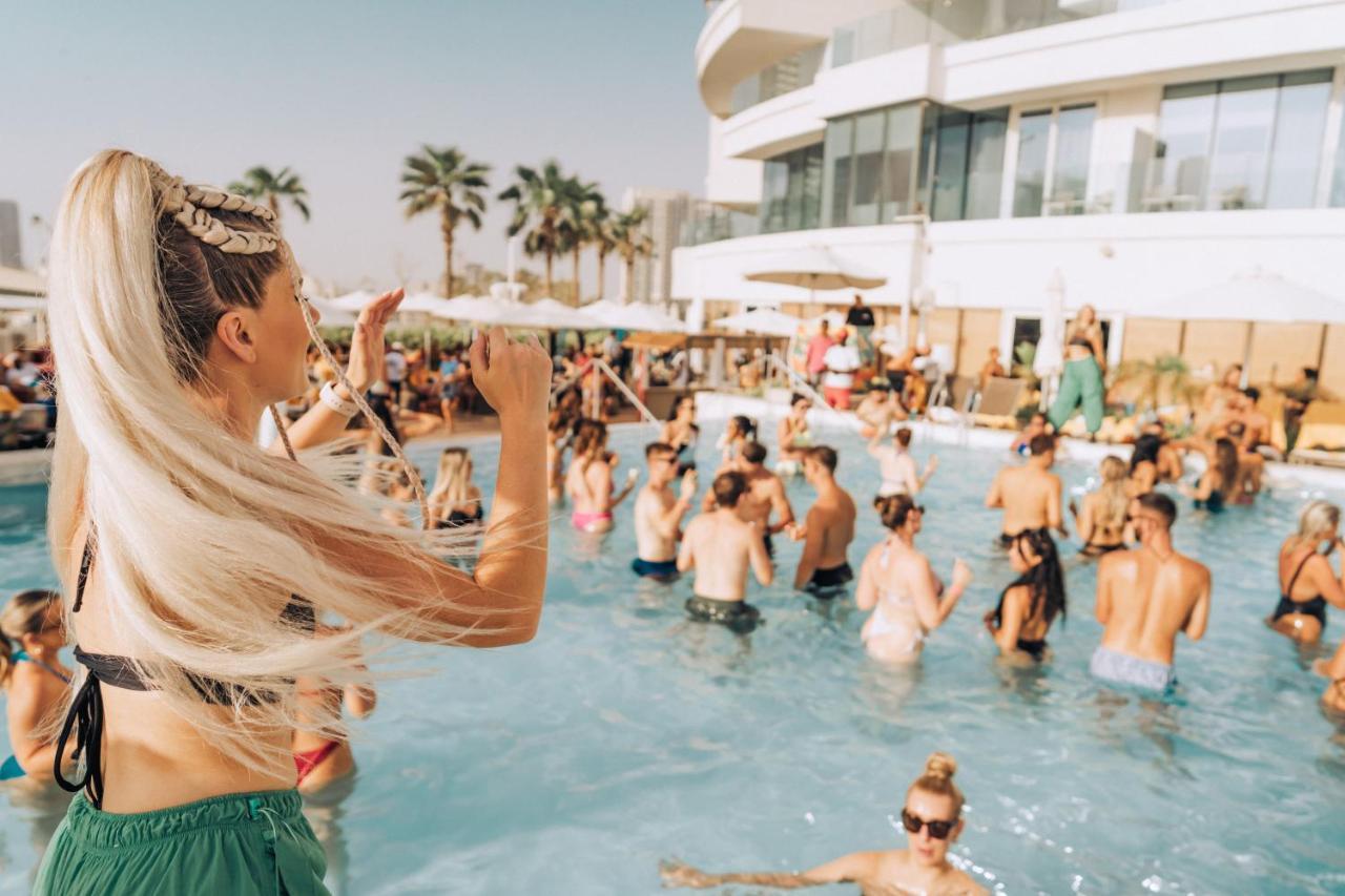 Five Jumeirah Village Дубай Экстерьер фото The photo features a lively pool party scene. In the foreground, a woman with long, blonde hair is seen raising her arm as if enjoying the moment. The pool is filled with many people, some swimming and others socializing. The atmosphere appears festi