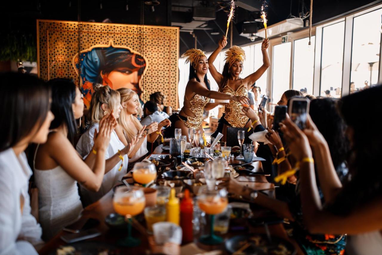 Five Jumeirah Village Дубай Экстерьер фото The photo depicts a lively restaurant scene where two performers are entertaining a group of seated diners. The performers, dressed in colorful and elaborate outfits, are holding sparklers, adding a festive atmosphere to the occasion. The diners, mos
