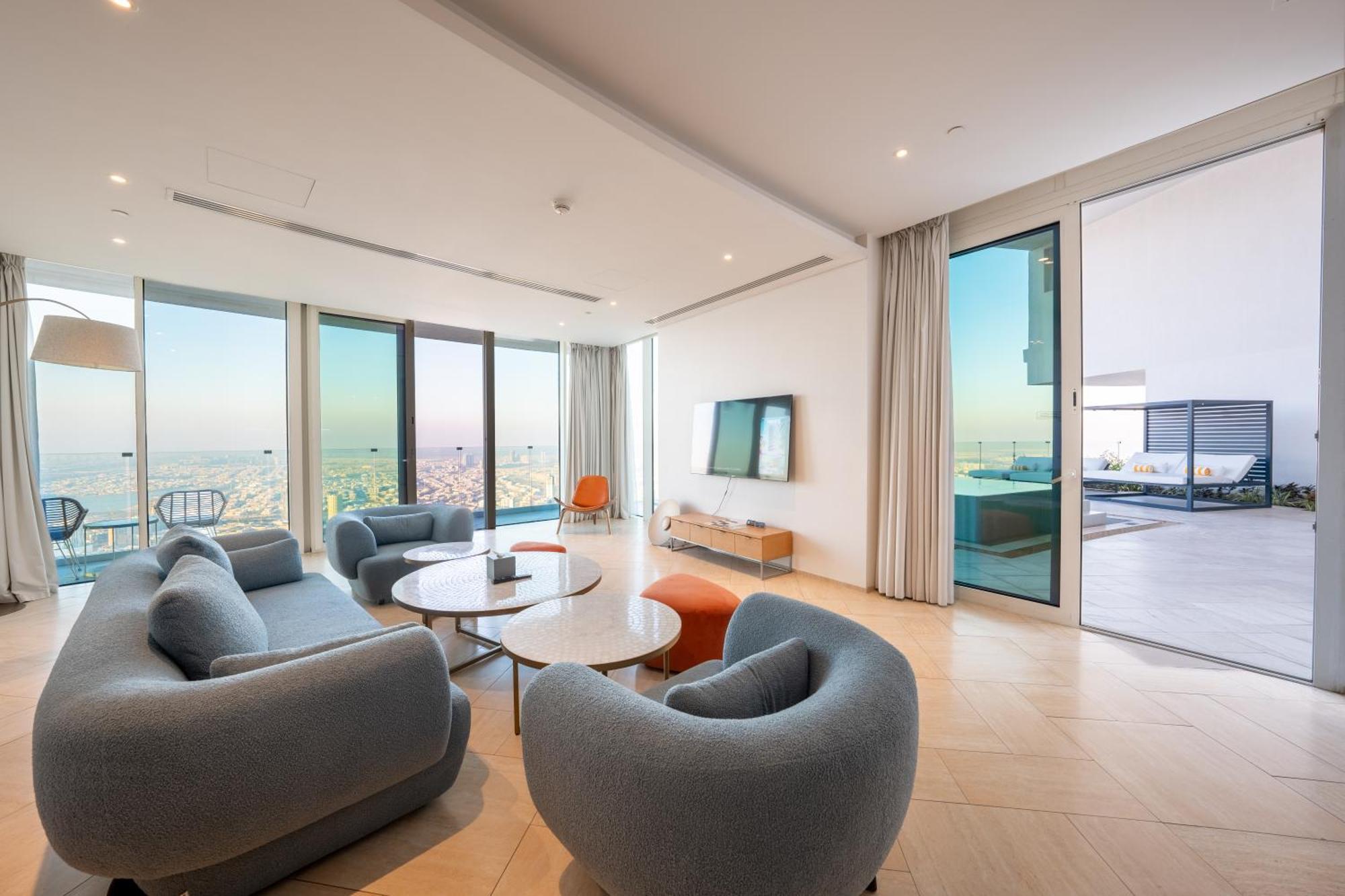 Five Jumeirah Village Дубай Экстерьер фото The photo shows a modern and stylish living room area with large windows that provide a scenic view. There are several rounded, gray sofas arranged around a couple of round coffee tables. A sleek television is mounted on the wall across from the seat