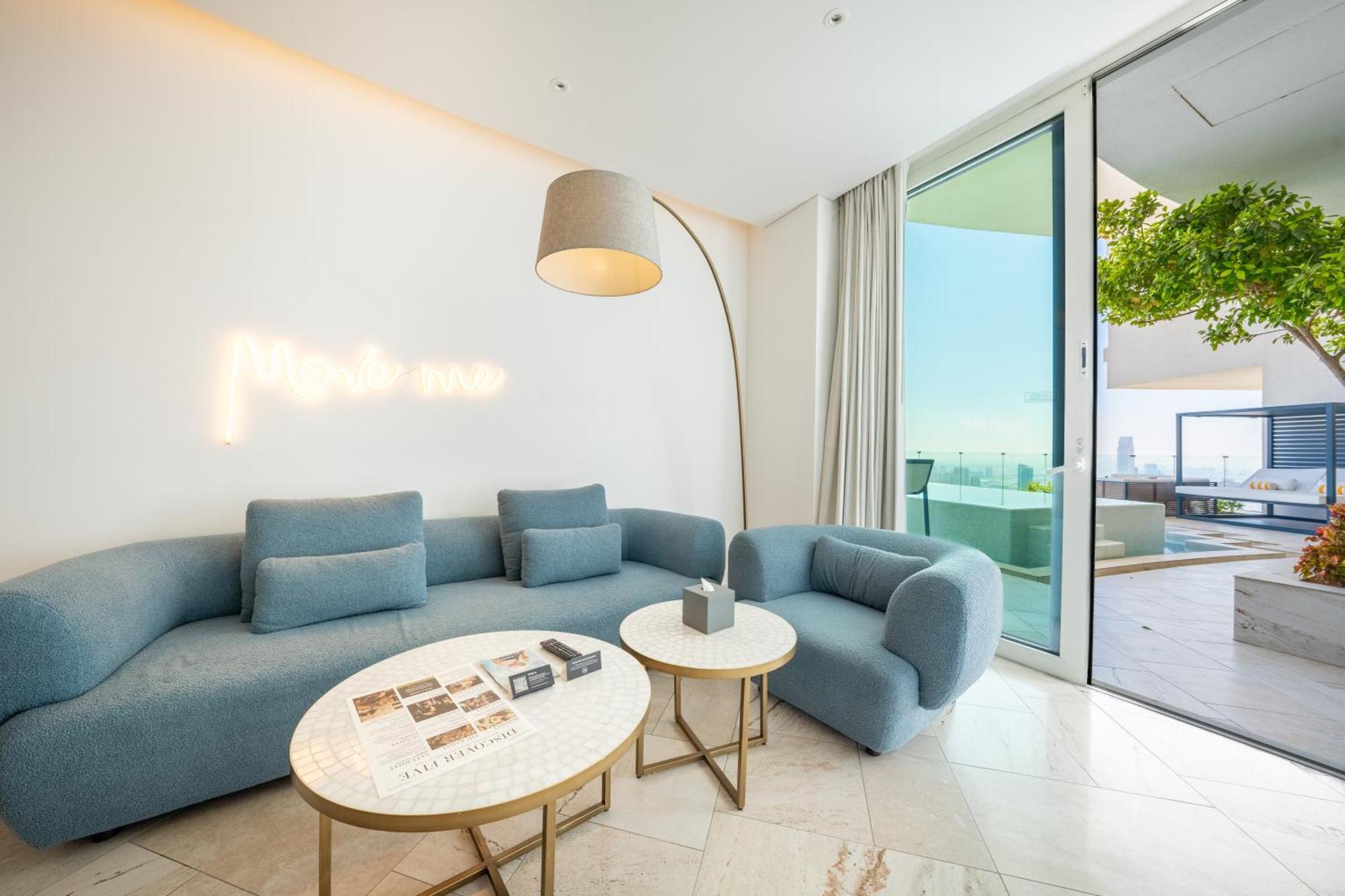 Five Jumeirah Village Дубай Экстерьер фото The photo shows a modern and stylish living space. It features a light blue sectional sofa and a matching armchair, creating a cozy seating area. Two round marble coffee tables are placed in front, one of which has some magazines or books on it. A ta