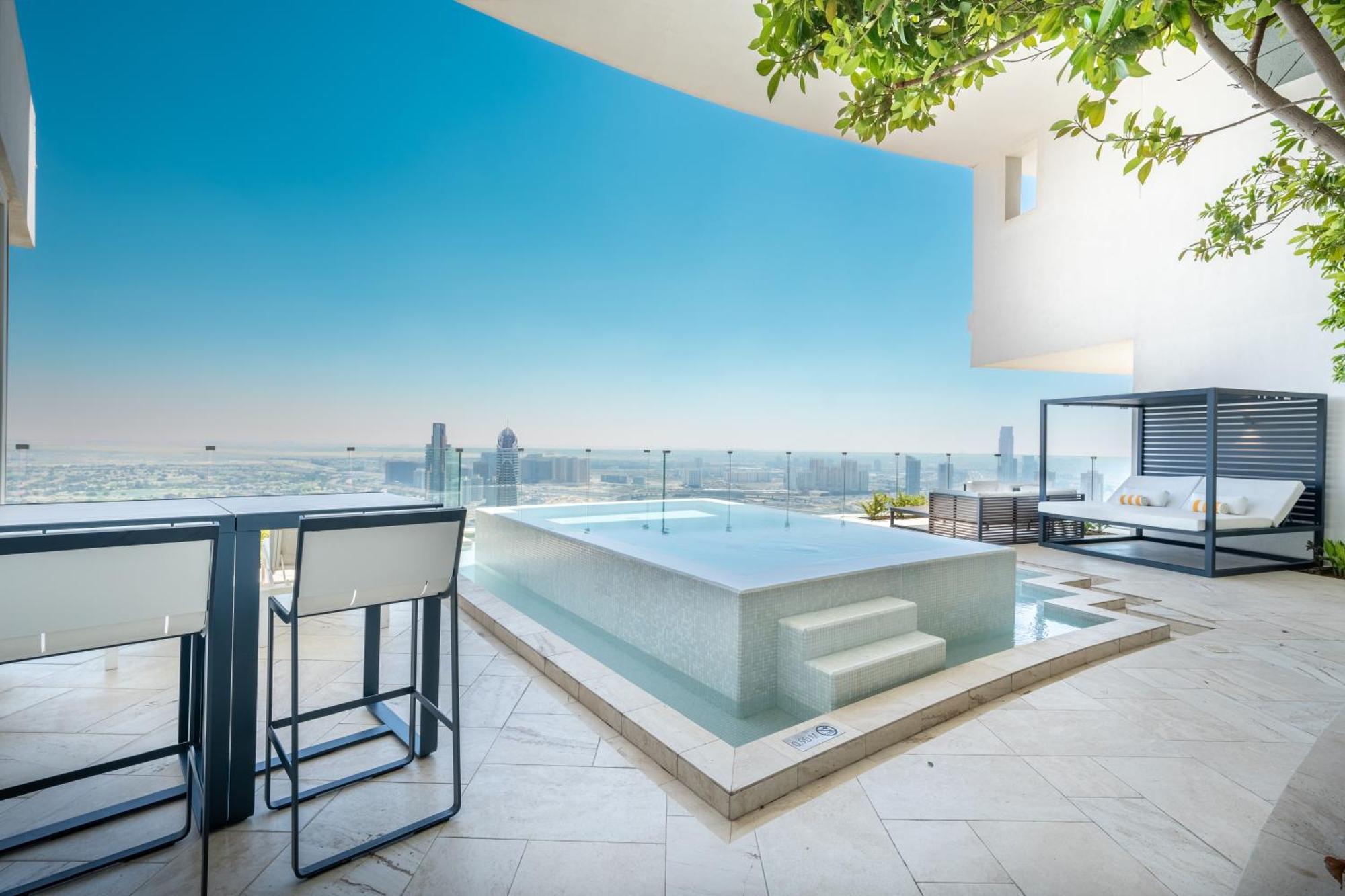 Five Jumeirah Village Дубай Экстерьер фото The photo shows a modern outdoor space featuring a sleek infinity pool. The pool appears to be situated on a terrace with a stunning view of a city skyline or landscape in the background. There are elegant lounge chairs and a shaded cabana area, crea