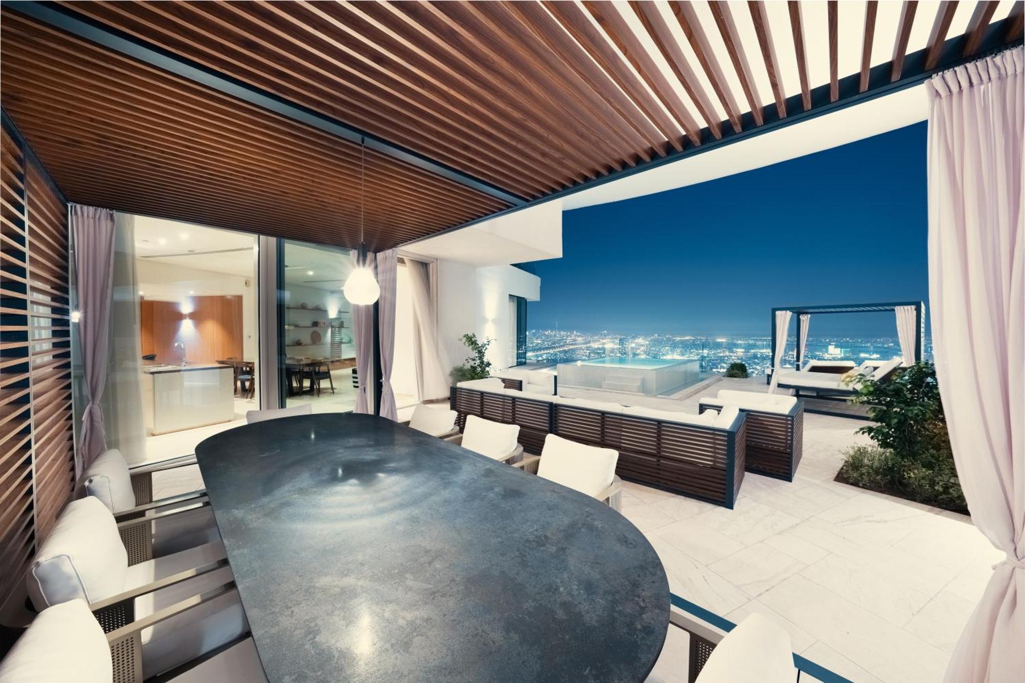 Five Jumeirah Village Дубай Экстерьер фото The photo shows a modern outdoor dining area with a sleek, curved table surrounded by several chairs. It features a stylish wooden ceiling with slats, creating a shaded atmosphere. In the background, large glass doors lead to the interior of a house,