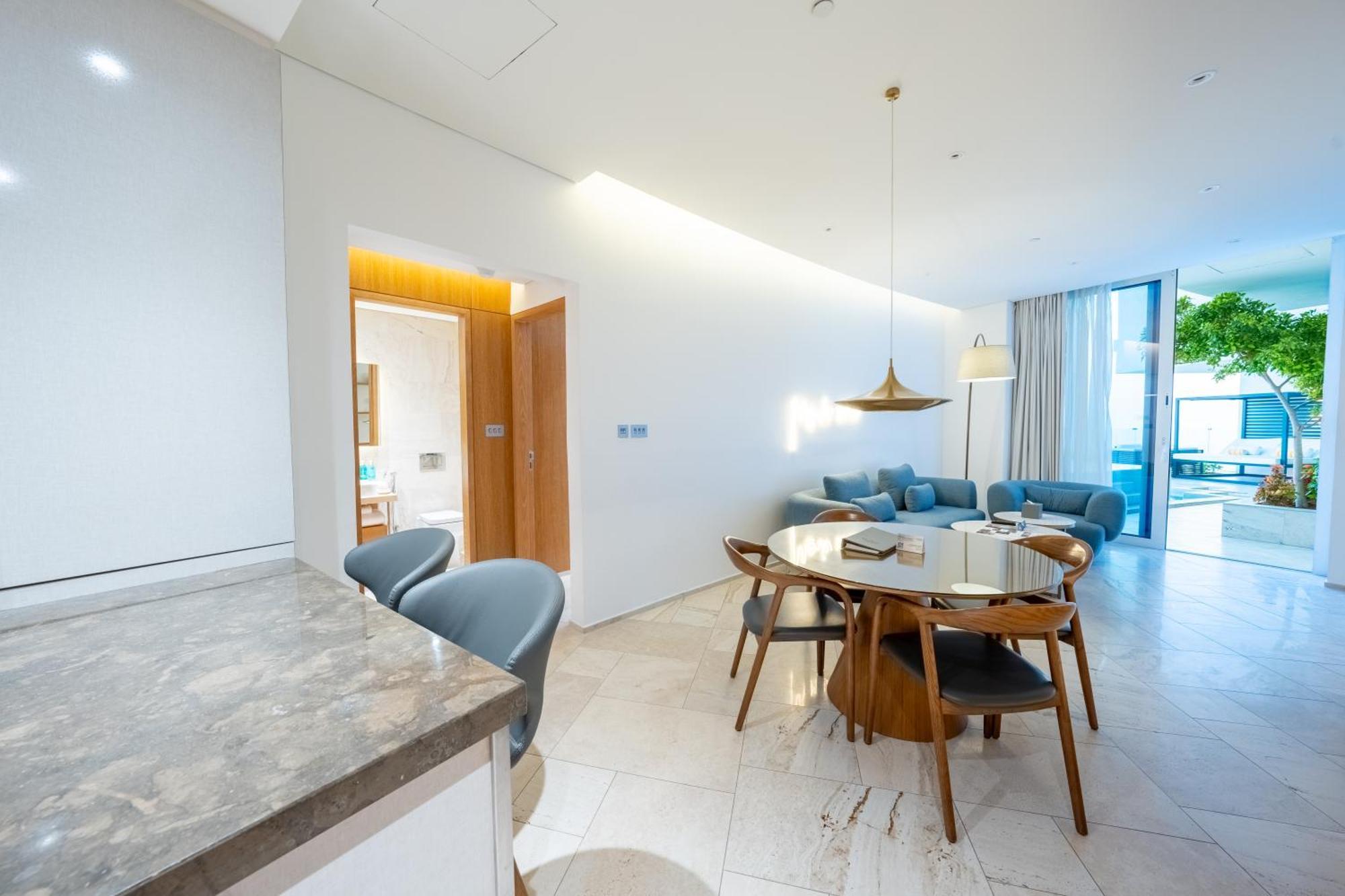 Five Jumeirah Village Дубай Экстерьер фото The photo shows a modern living space that combines a dining area and a lounging area. On the left, there is a kitchen counter with a marble surface and several gray chairs around it. In the center, there is a round dining table made of wood with sev