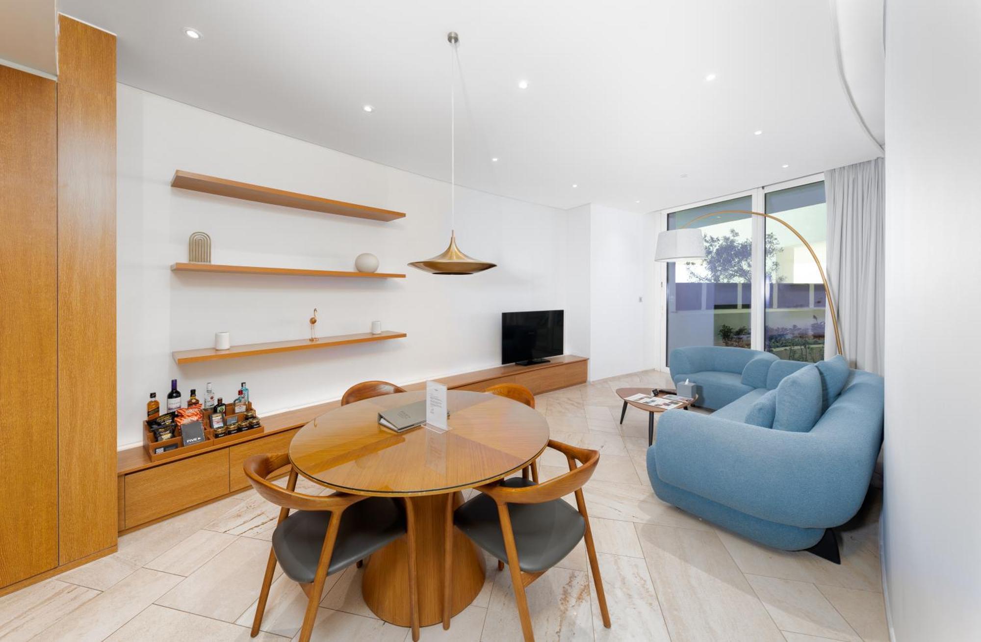Five Jumeirah Village Дубай Экстерьер фото The image shows a modern living space that features a cozy and stylish design. On one side, there is a round wooden dining table surrounded by four chairs. The table is set with a small item, possibly a decorative piece or a book. 

A pendant light h