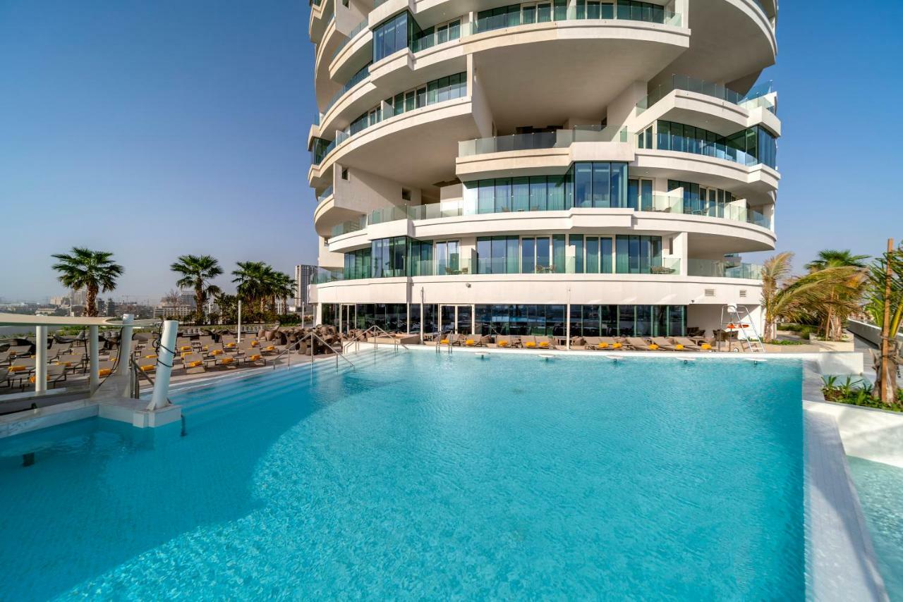 Five Jumeirah Village Дубай Экстерьер фото The photo depicts a modern building with a distinctive architectural design, featuring multiple curved balconies. In front of the building, there is a large, inviting swimming pool with clear, turquoise water. Surrounding the pool are loungers for re